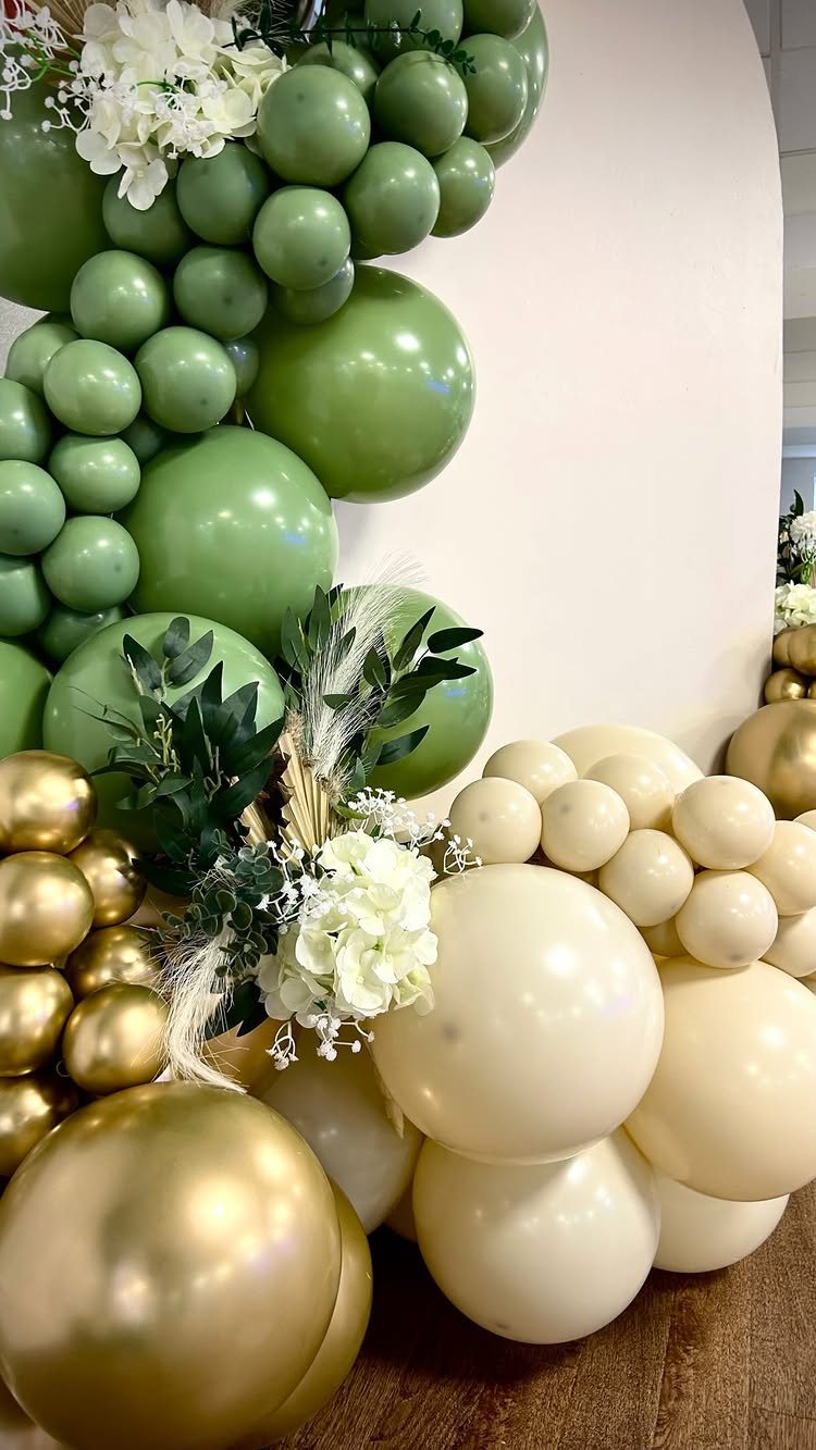 Green, cream, and gold balloons decorated with white flowers and green leaves against a white wall.