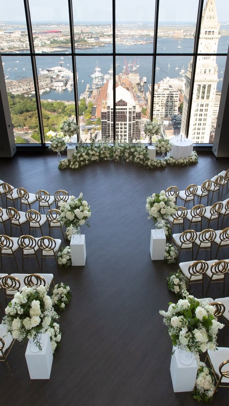 White floral arrangements and gold chairs arranged for a wedding ceremony overlooking a city harbor.