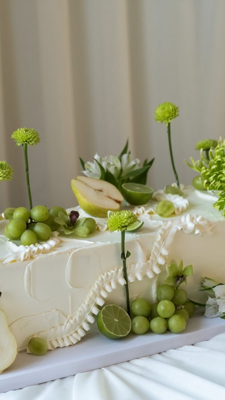 White frosted cake decorated with green grapes, lime slices, pear slices, and green flowers.