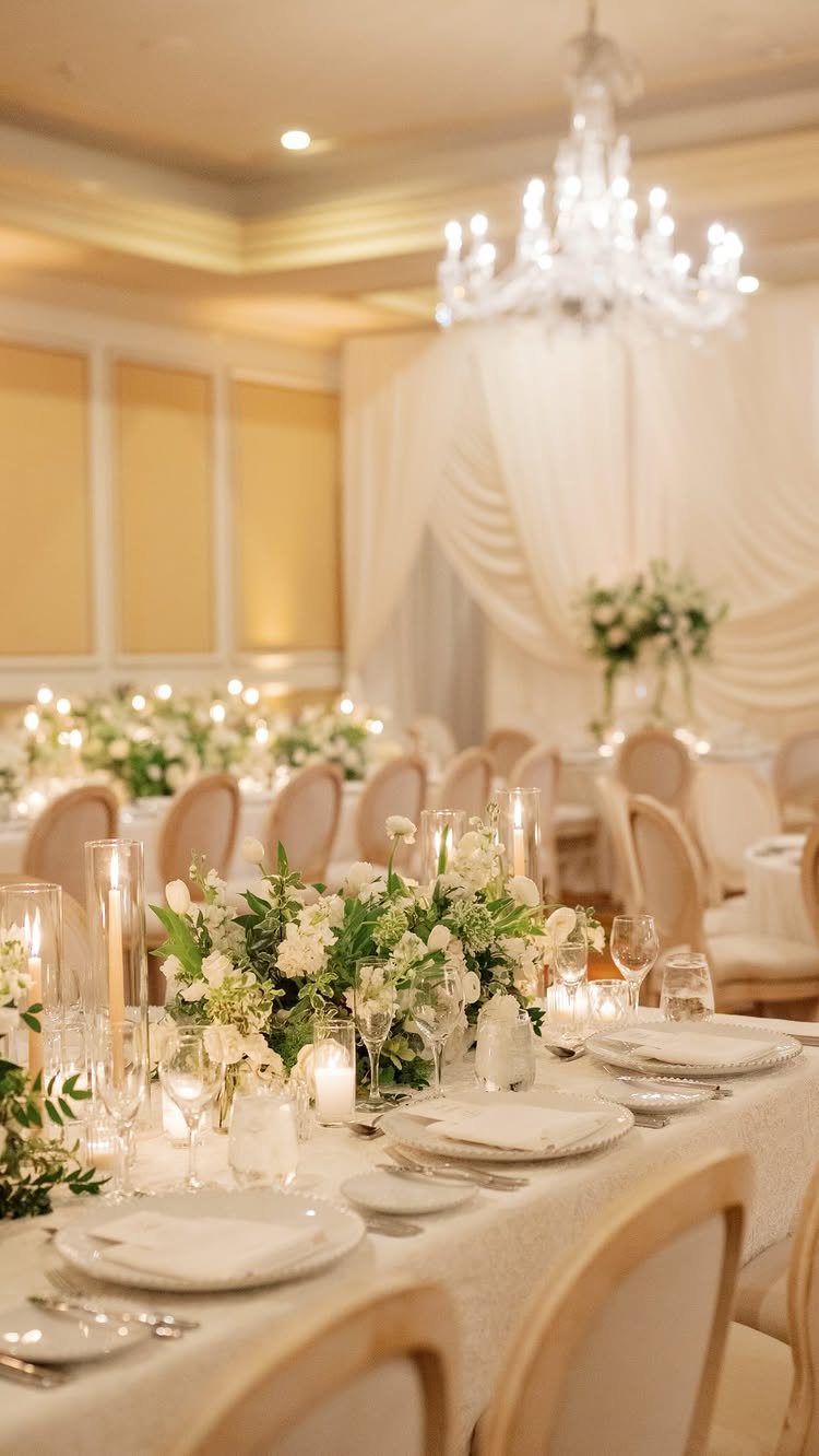 White wedding tablescapes with greenery centerpieces and a large crystal chandelier in a formal ballroom.