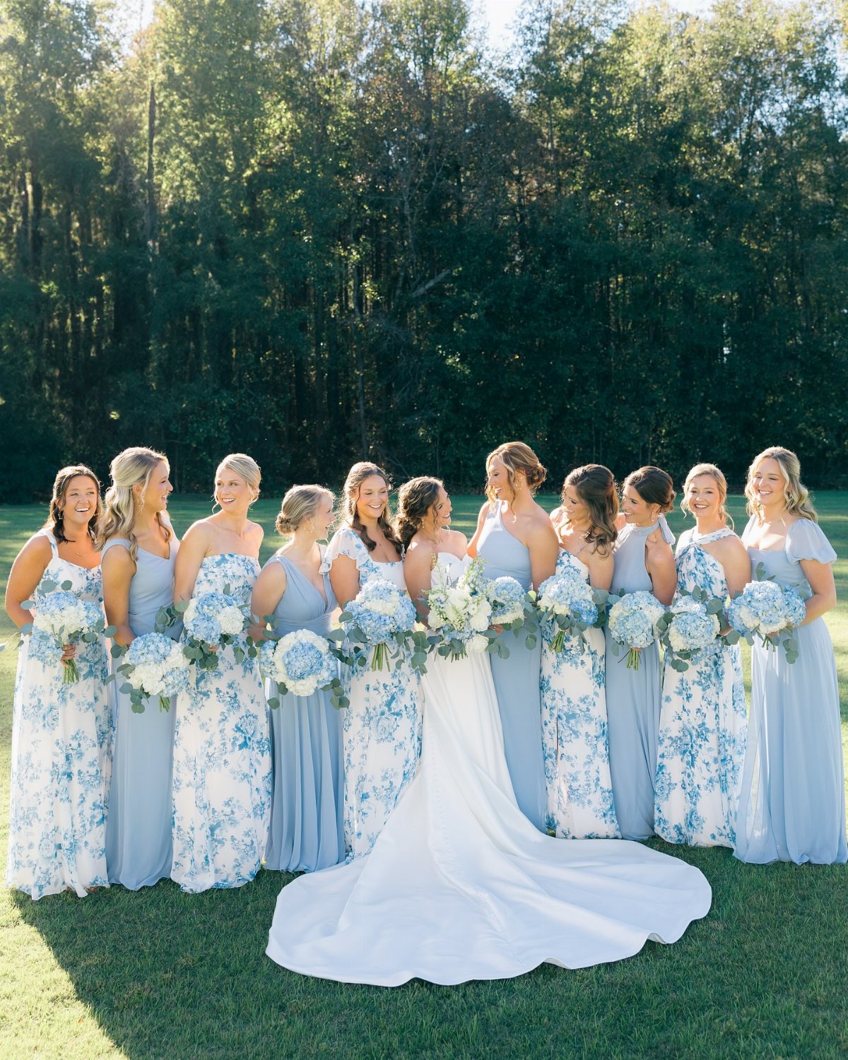 Bridesmaids in blue floral dresses holding white and blue bouquets standing with a bride outdoors.
