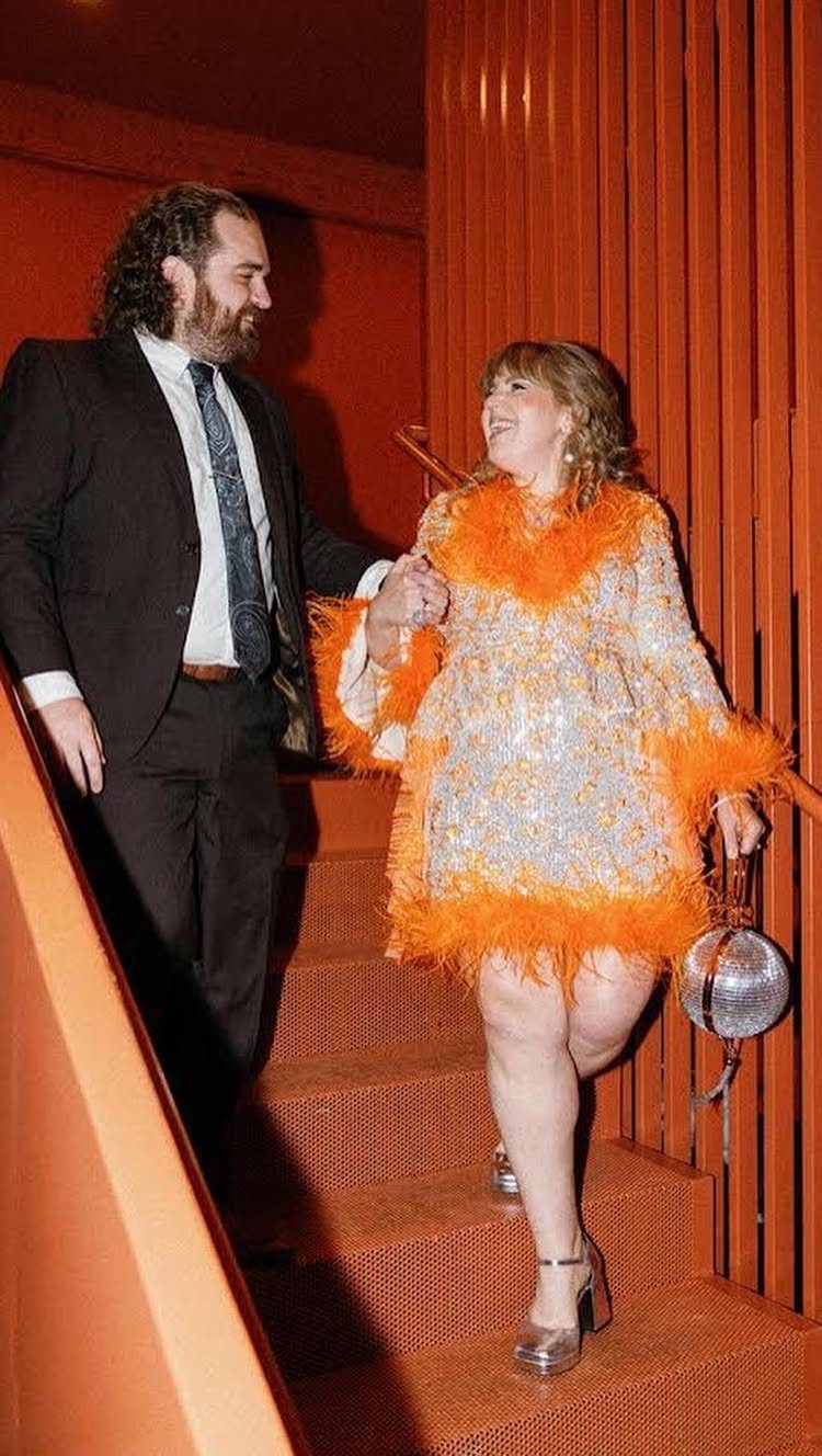 A woman in a sparkly silver and orange feathered dress walks down stairs with a man.
