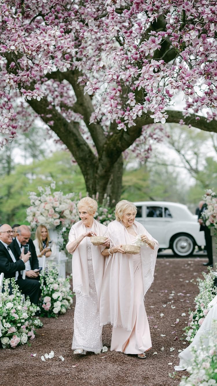 Two elderly women in pink dresses walk down an aisle carrying flower petal baskets.