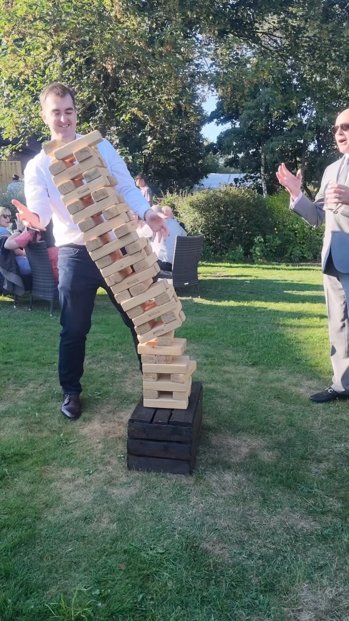 A large wooden block tower tumbling down on a grassy lawn during an outdoor event.