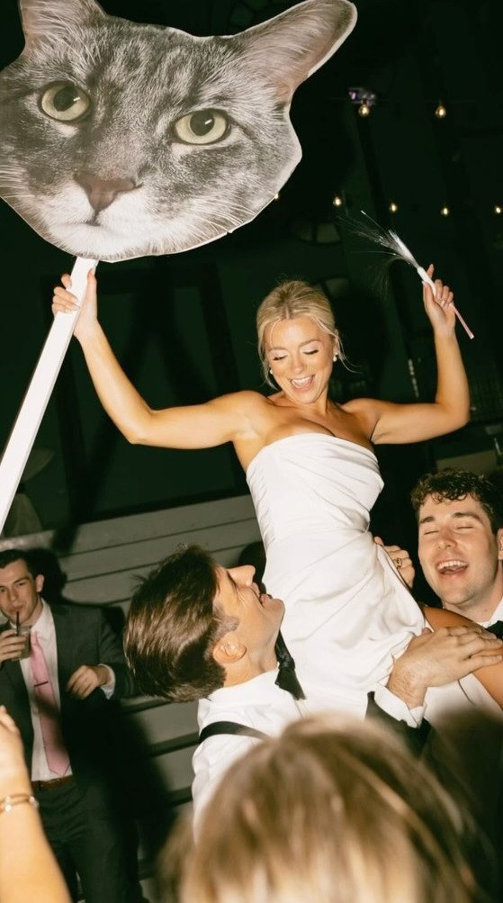 A bride holding a large cat head cutout while being lifted on shoulders during a wedding celebration.