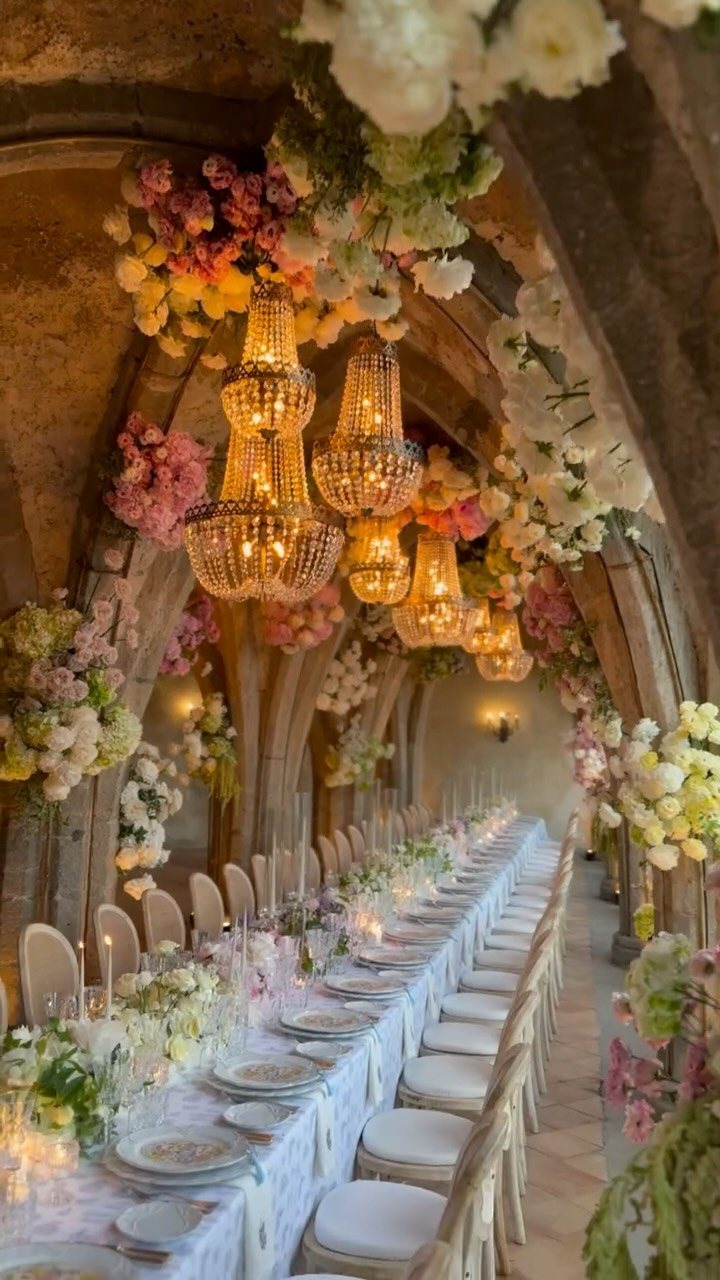 Long banquet table set under stone arches with crystal chandeliers and floral arrangements.