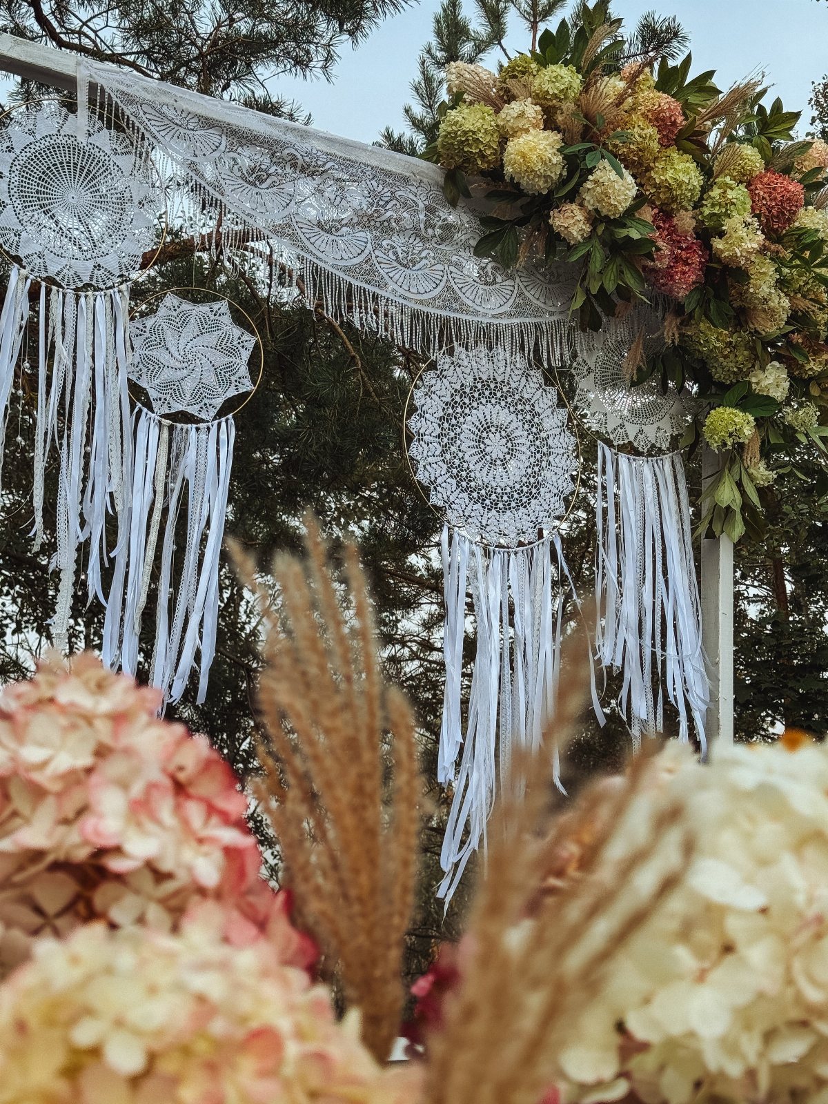 White wedding arch featuring large lace dreamcatchers, hanging ribbons, and colorful floral arrangements.