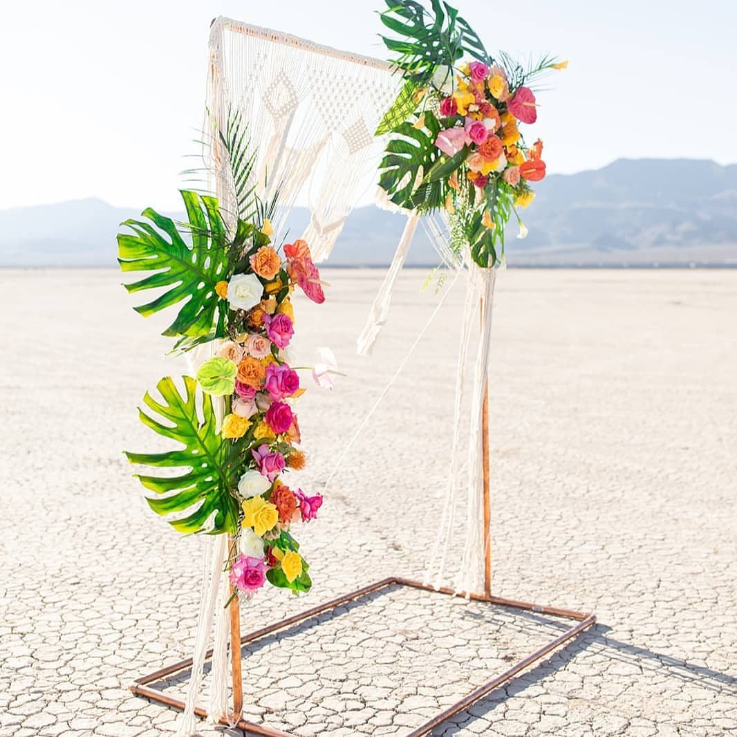 Copper pipe wedding arch decorated with tropical leaves, colorful flowers, and white macrame in a desert.