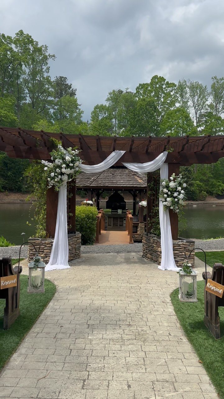 Dark wood pergola decorated with white fabric and large white floral arrangements by a lake.