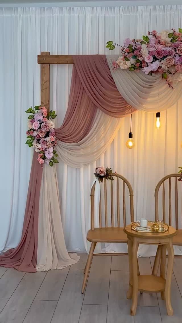 Wooden arch with draped mauve and cream fabric and pink flowers against white curtains.