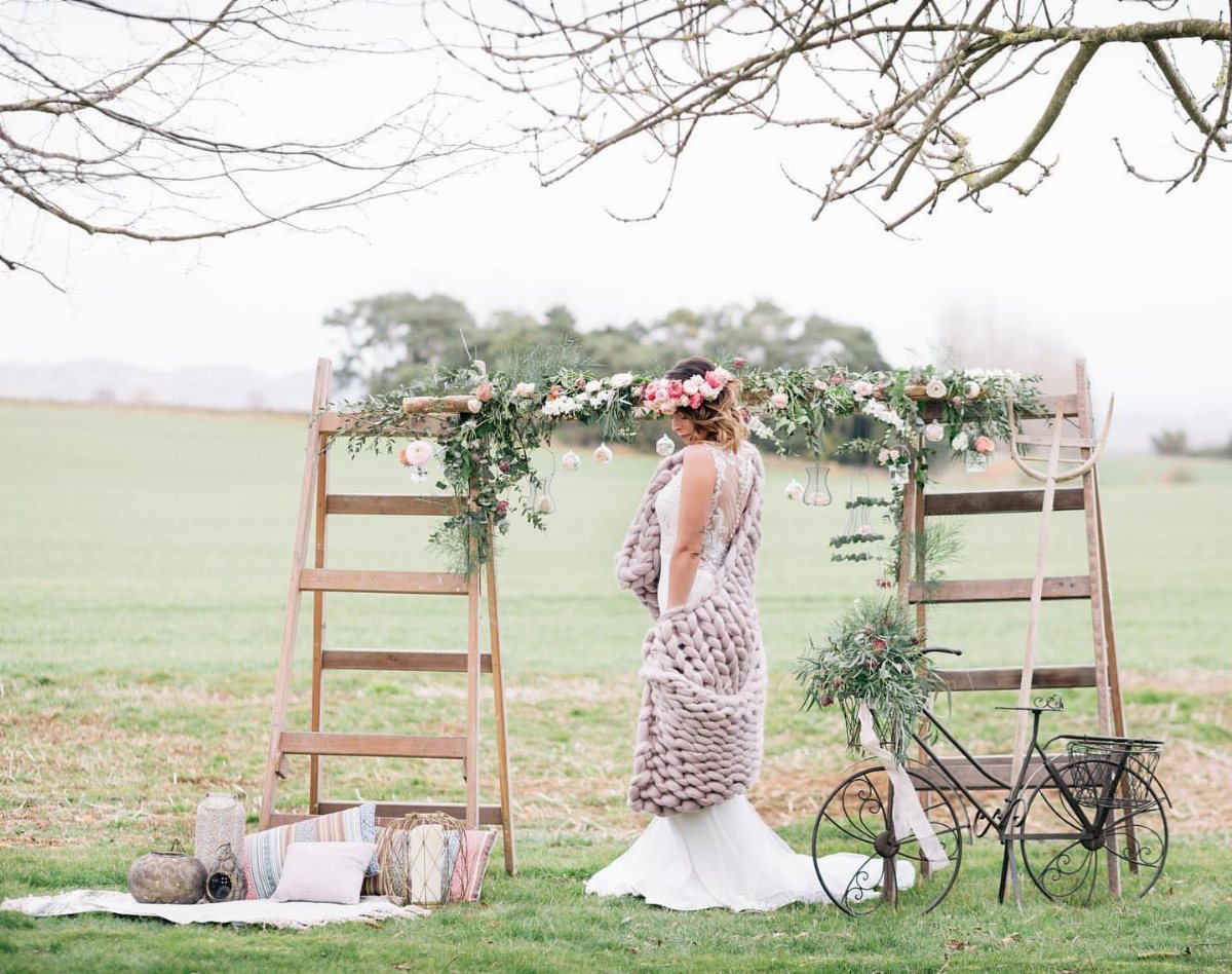 Two wooden ladders decorated with greenery and flowers serve as a wedding arch in a field.