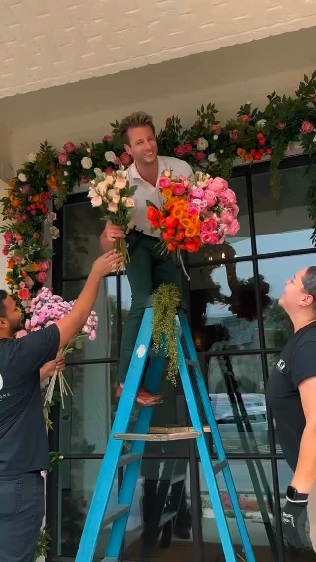 Men decorating a large glass door with a vibrant floral garland using a blue ladder.