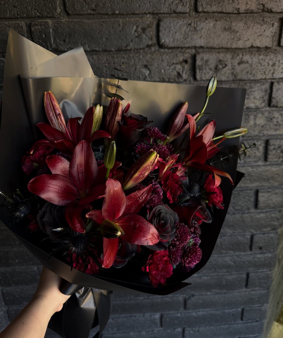A hand holding a bouquet of dark red lilies and black flowers wrapped in black paper.