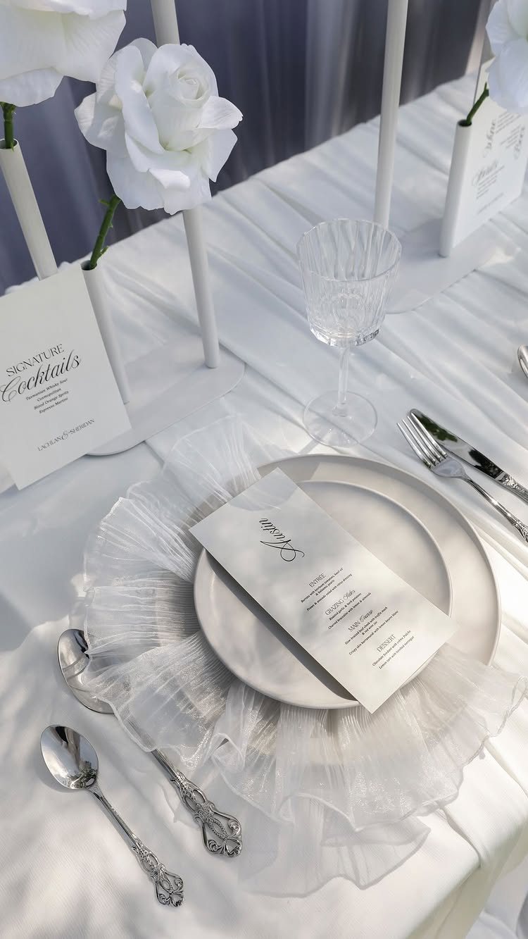 White wedding table setting with ruffled tulle charger, silver cutlery, menu, and white roses.