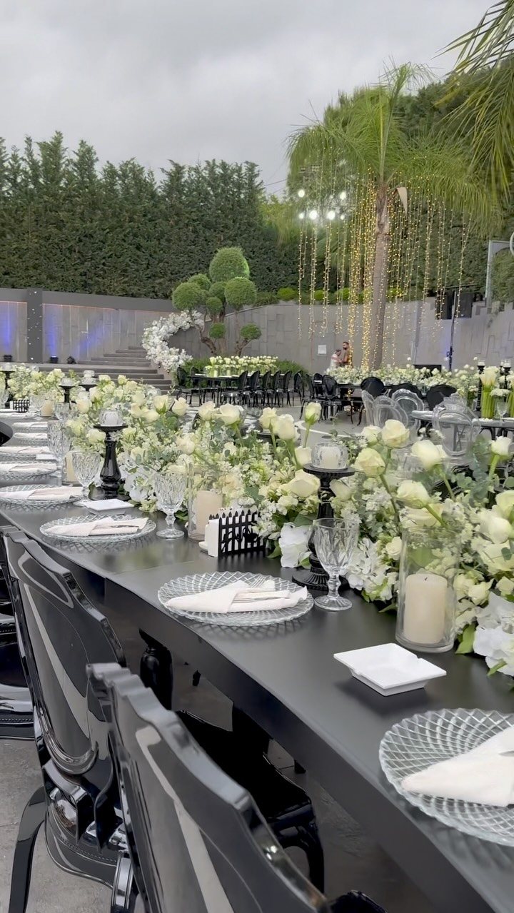 Long black wedding table featuring white rose centerpieces, candles, and elegant glassware in an outdoor setting.