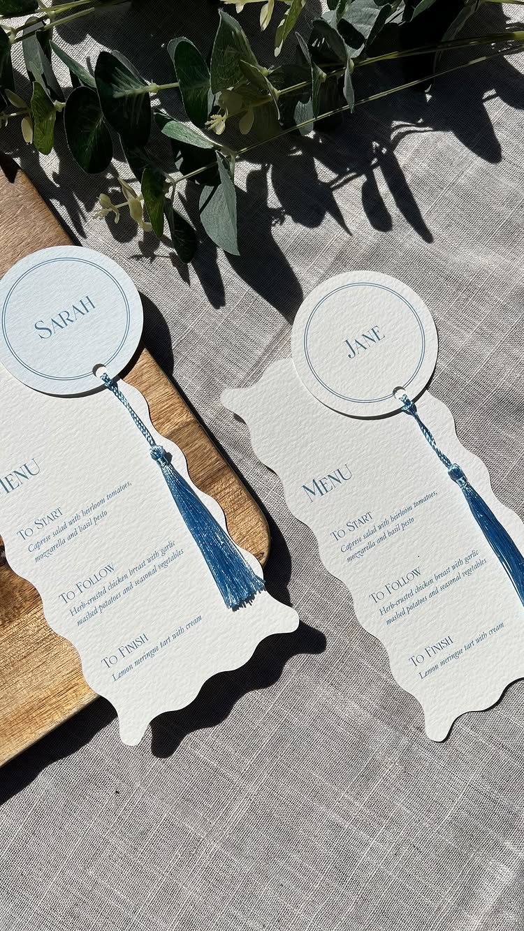 Two scalloped menu cards with blue tassel name tags resting on a linen tablecloth with greenery.