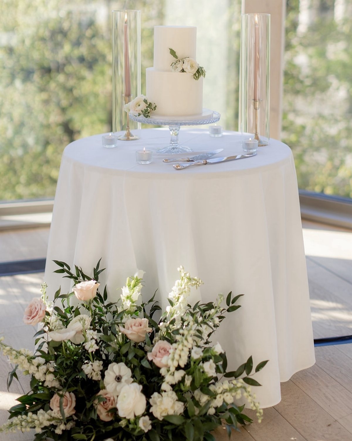 A white two-tier wedding cake with flowers on a round table with white linens.