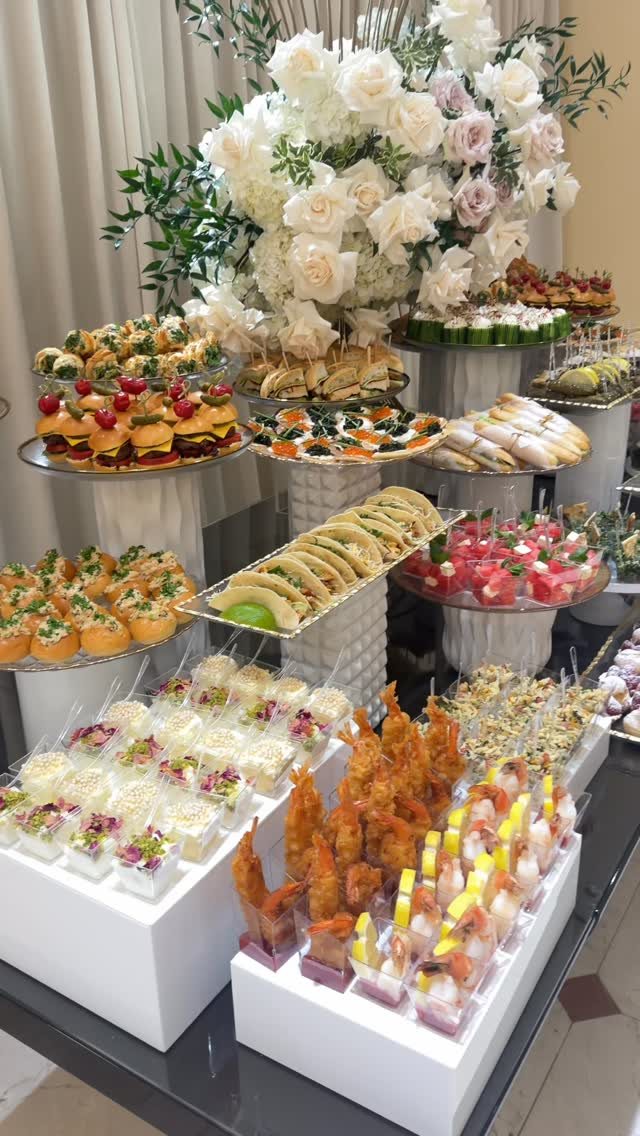 Assorted appetizers and mini sliders arranged on white tiered stands with large white flowers.