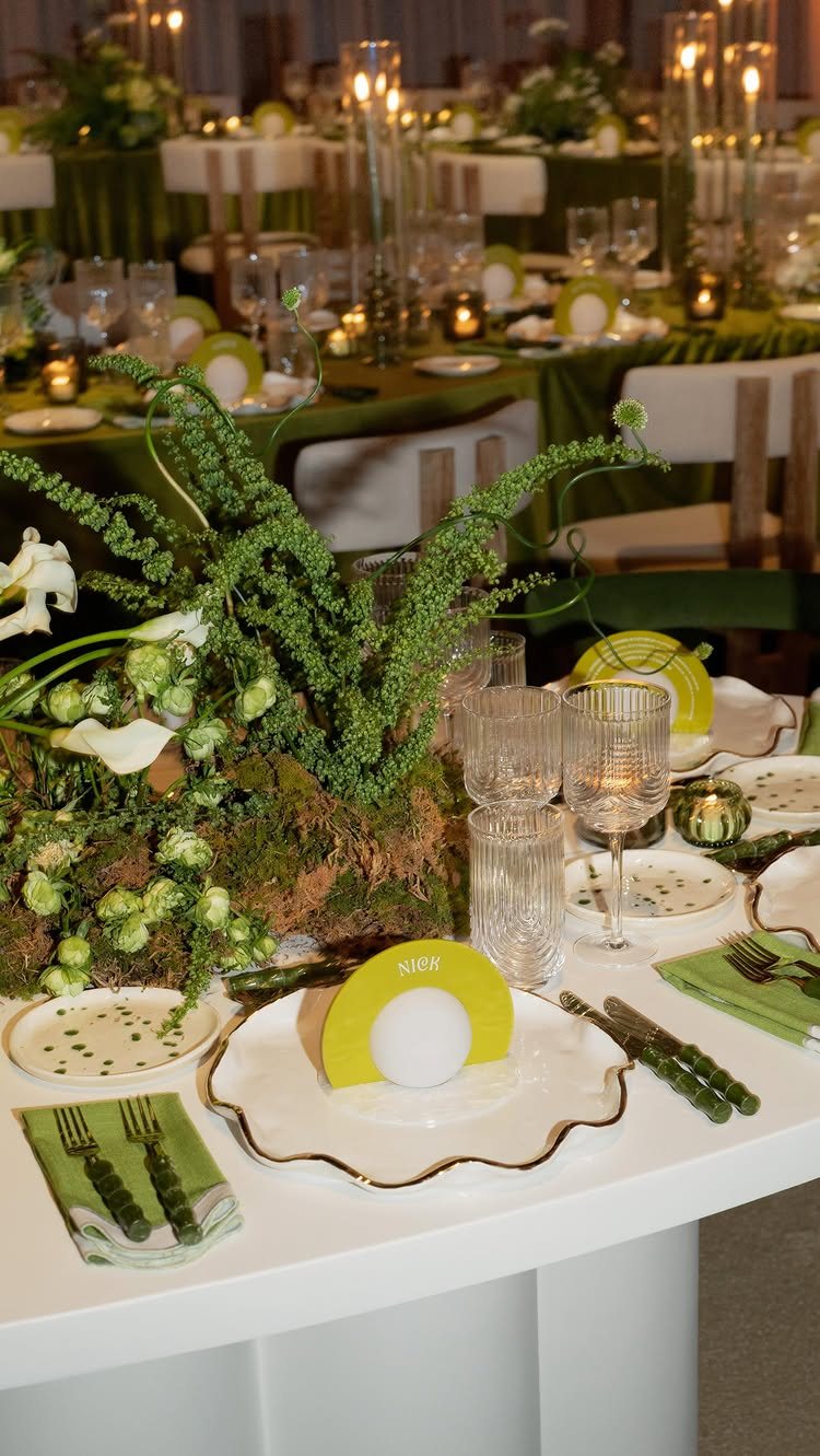 A white wedding reception table set with green accents, crystal glassware, and a mossy floral centerpiece.