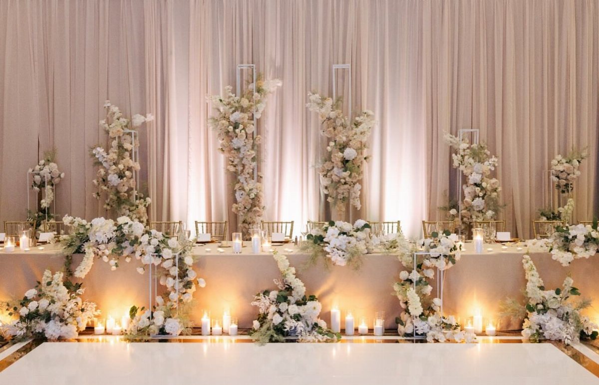 A long wedding head table decorated with lush white floral arrangements and glowing candles.