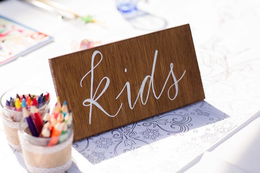 Wooden sign reading Kids next to jars of colored pencils and coloring books on a white table.