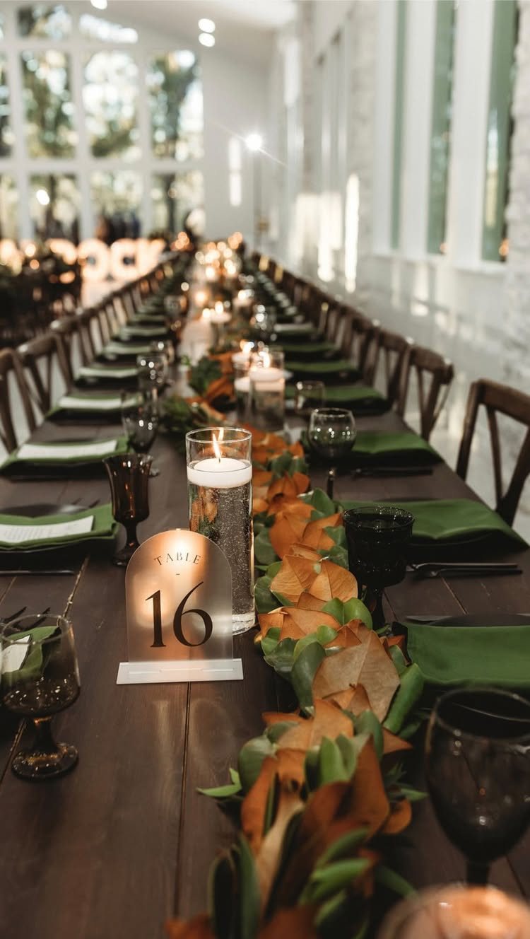 Long wooden dining table decorated with green napkins, dark glassware, and an autumnal leaf garland.