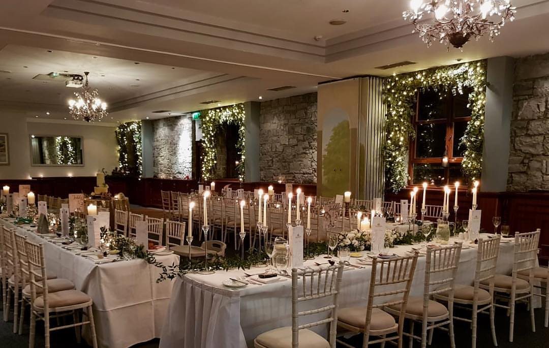U-shaped dining tables decorated with white linens, greenery, and many tall taper candles in a banquet hall.