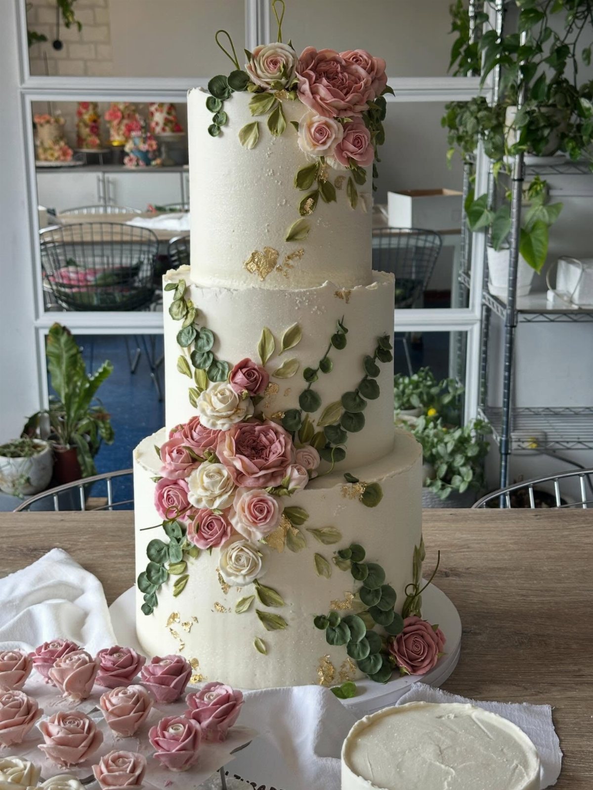 Three-tiered white wedding cake decorated with pink roses, green leaves, and gold leaf accents.