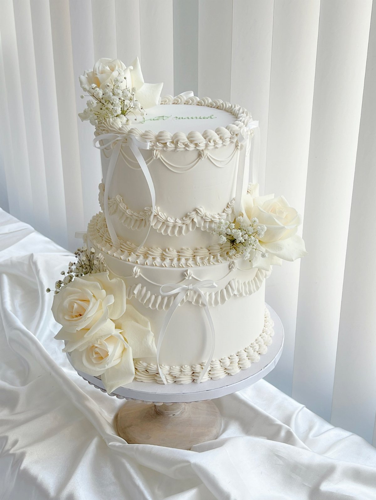 Two-tier white wedding cake with buttercream piping, white ribbons, and fresh white roses.