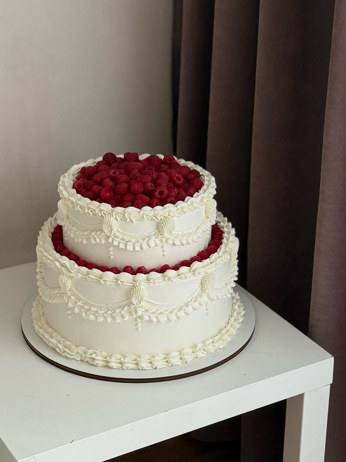 Two-tier white buttercream wedding cake with intricate piped lace details and fresh red raspberries on top.