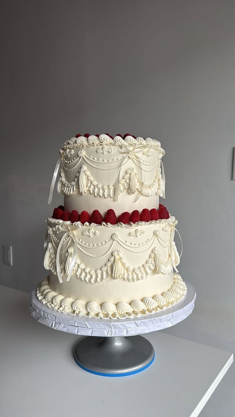 Two-tier white buttercream cake with vintage piped decorations and fresh red raspberries on a silver stand.