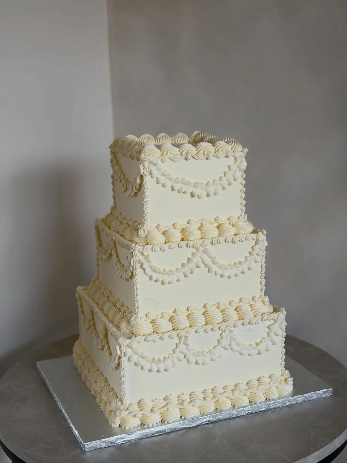 Three-tiered square wedding cake with cream buttercream and intricate piped lace decorations.