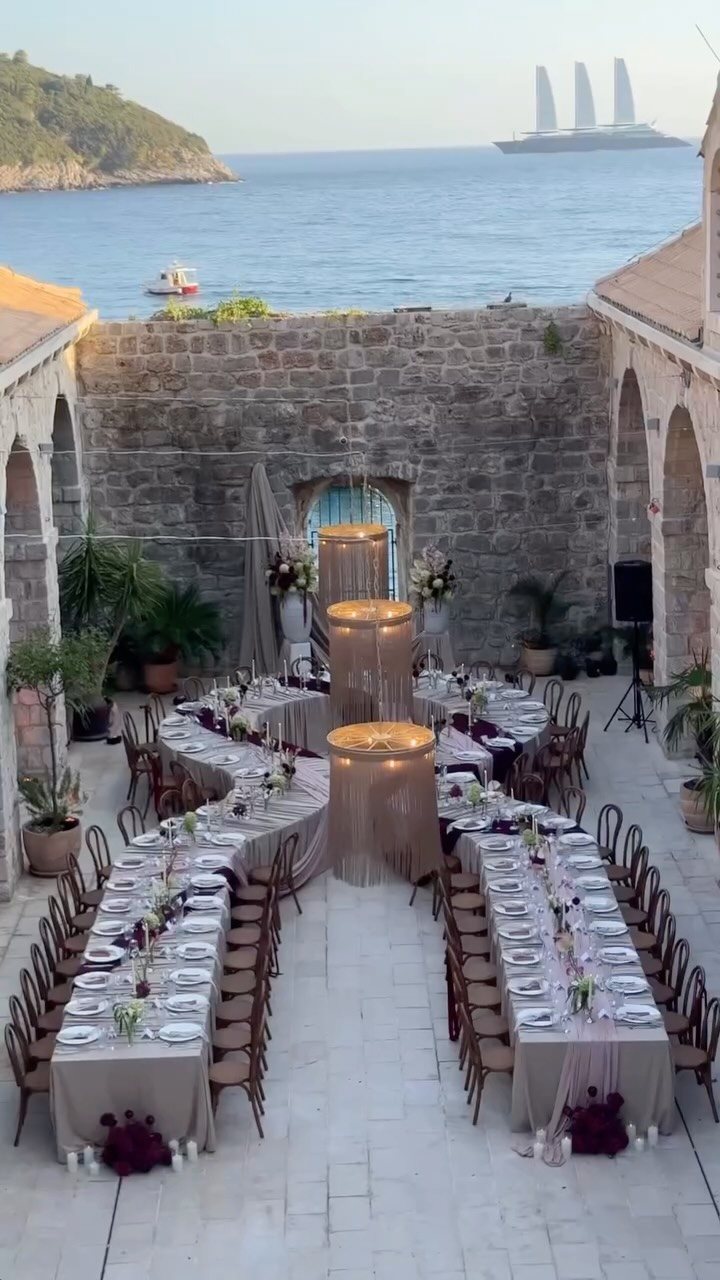 Long banquet tables set for a formal dinner on a stone patio overlooking the sea.