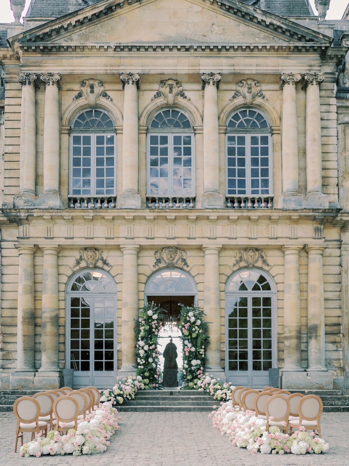 Floral arrangements and chairs line an aisle leading to an archway in front of a grand building.