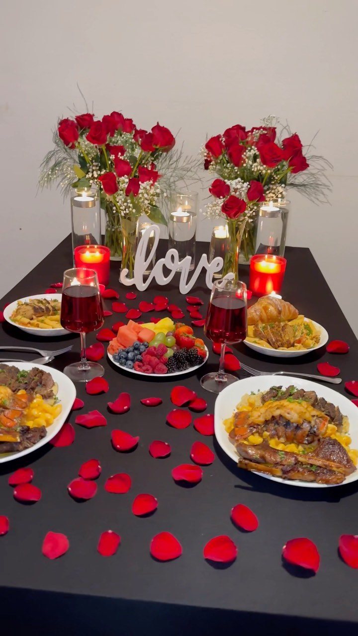 A romantic dinner table set with red roses, candles, wine, and various food dishes.