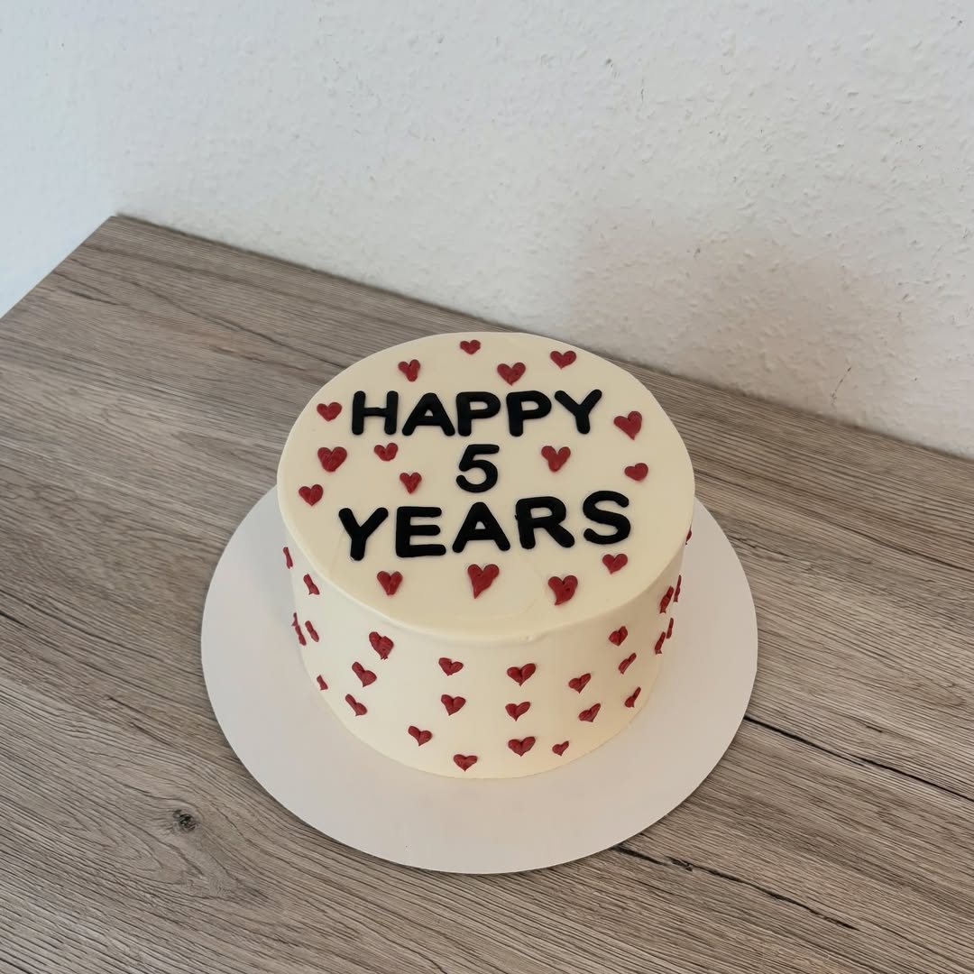 A white frosted cake decorated with small red hearts and black text saying Happy 5 Years.
