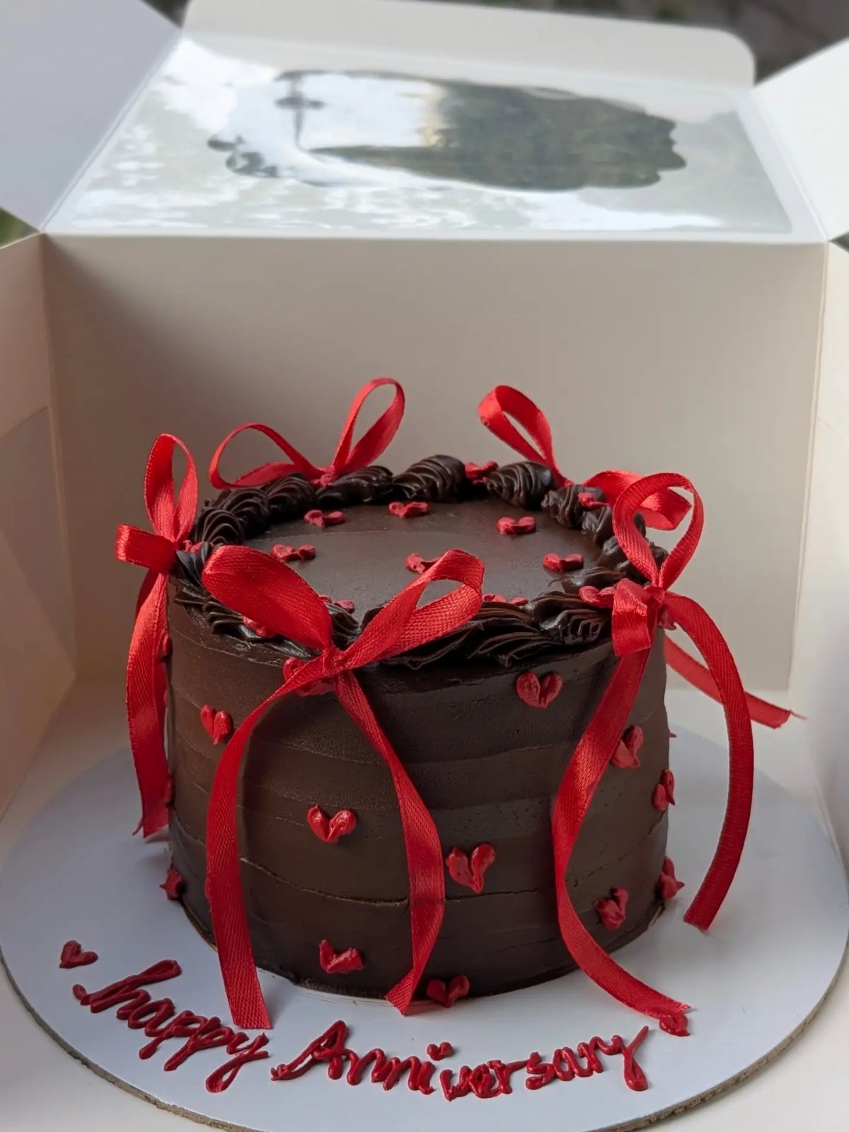 Chocolate cake decorated with red frosting hearts and large red ribbons in a white box.