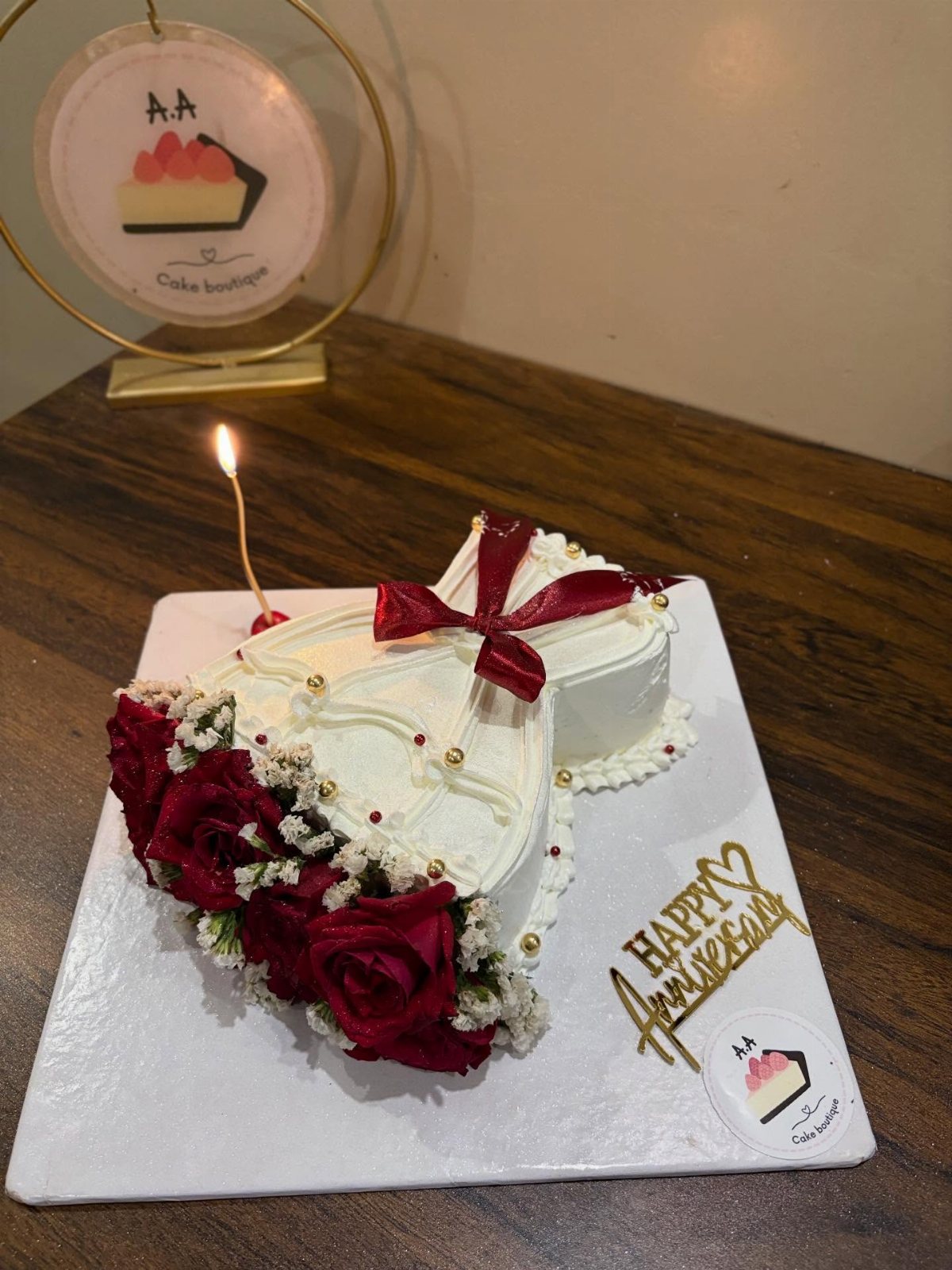 Heart-shaped white cake decorated with red roses, white flowers, and a gold anniversary topper on a wooden table.