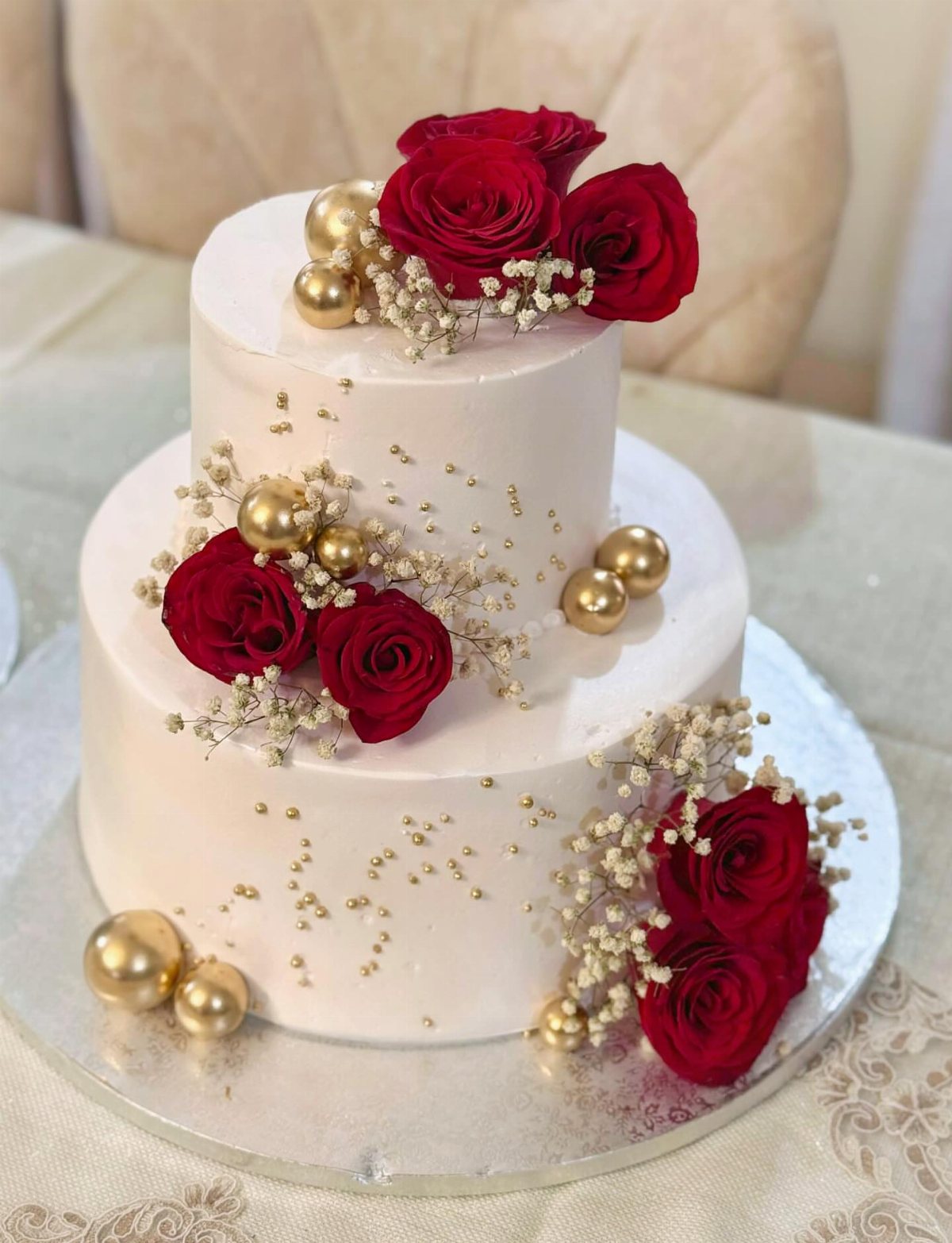 Two-tiered white cake decorated with red roses, gold spheres, and delicate white baby's breath flowers.