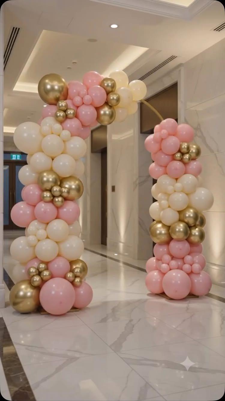 A large archway made of pink, cream, and gold balloons in a marble hallway.