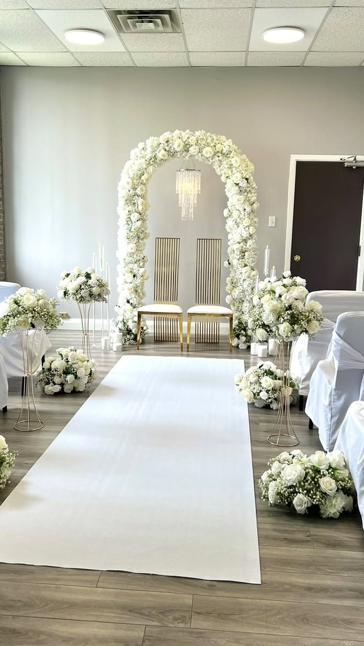 White floral wedding arch with a crystal chandelier and gold chairs over a white aisle runner.