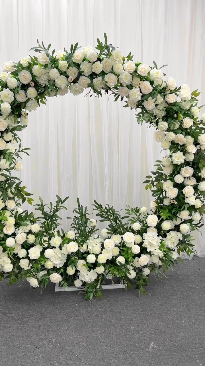 A circular wedding arch decorated with white roses and green leaves against white drapes.