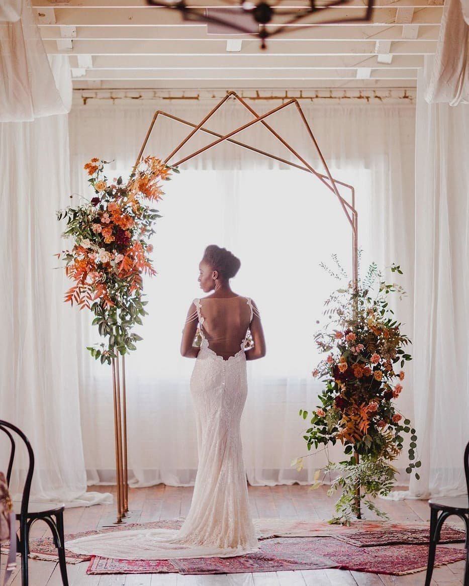 Woman in white dress standing before a geometric copper wedding arch with floral arrangements.