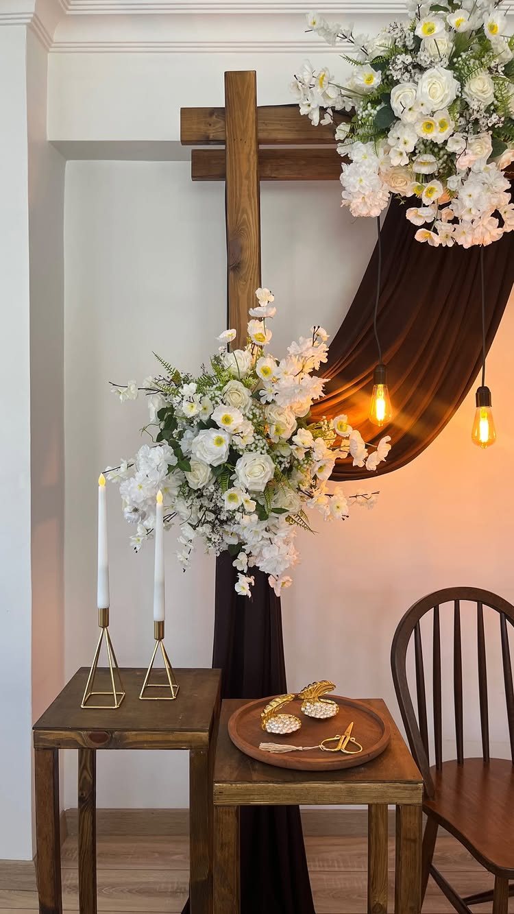 Wooden cross structure decorated with white flowers, brown fabric, and hanging Edison bulbs.