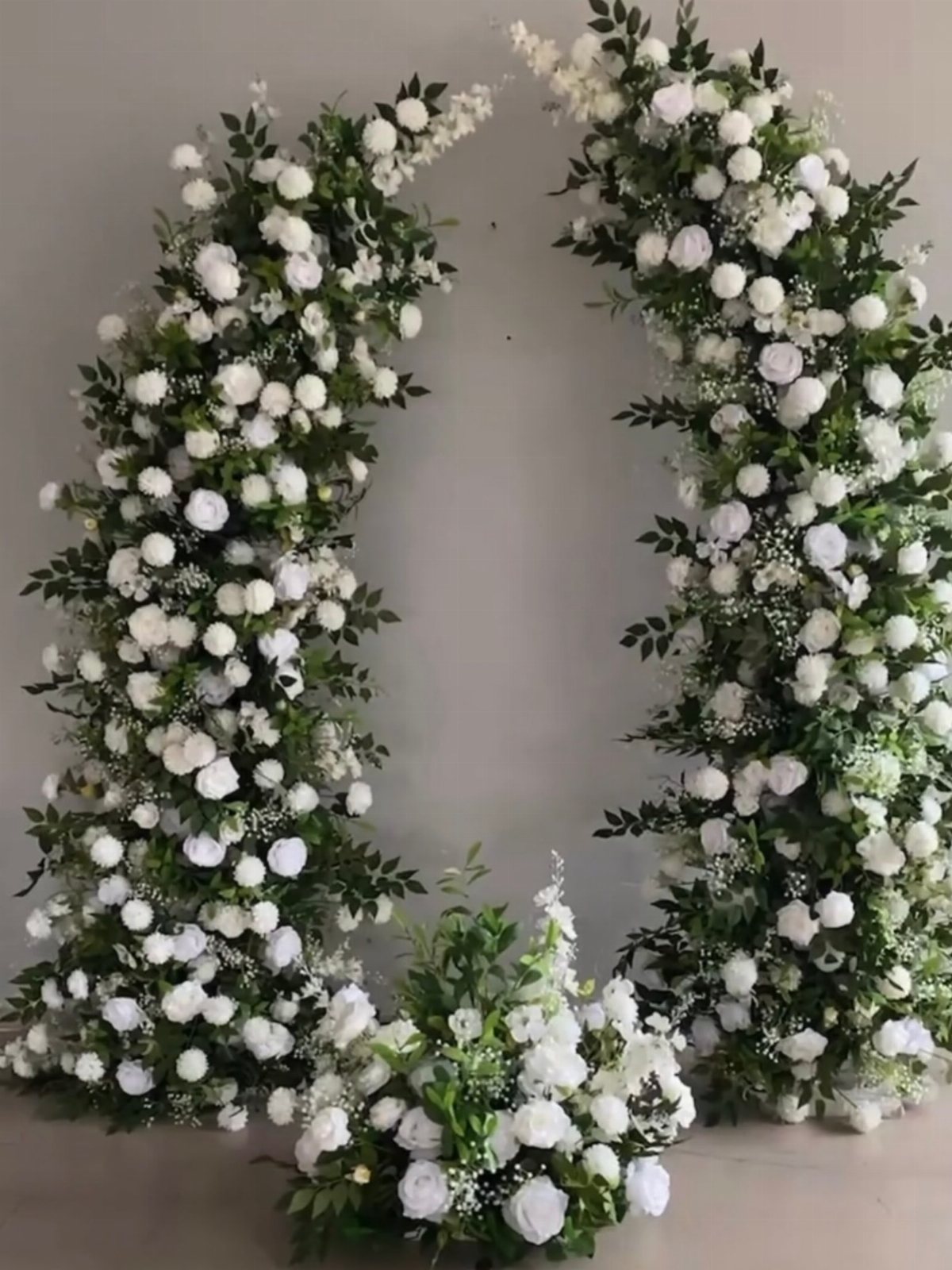 A large circular wreath made of white roses and green foliage against a grey wall.