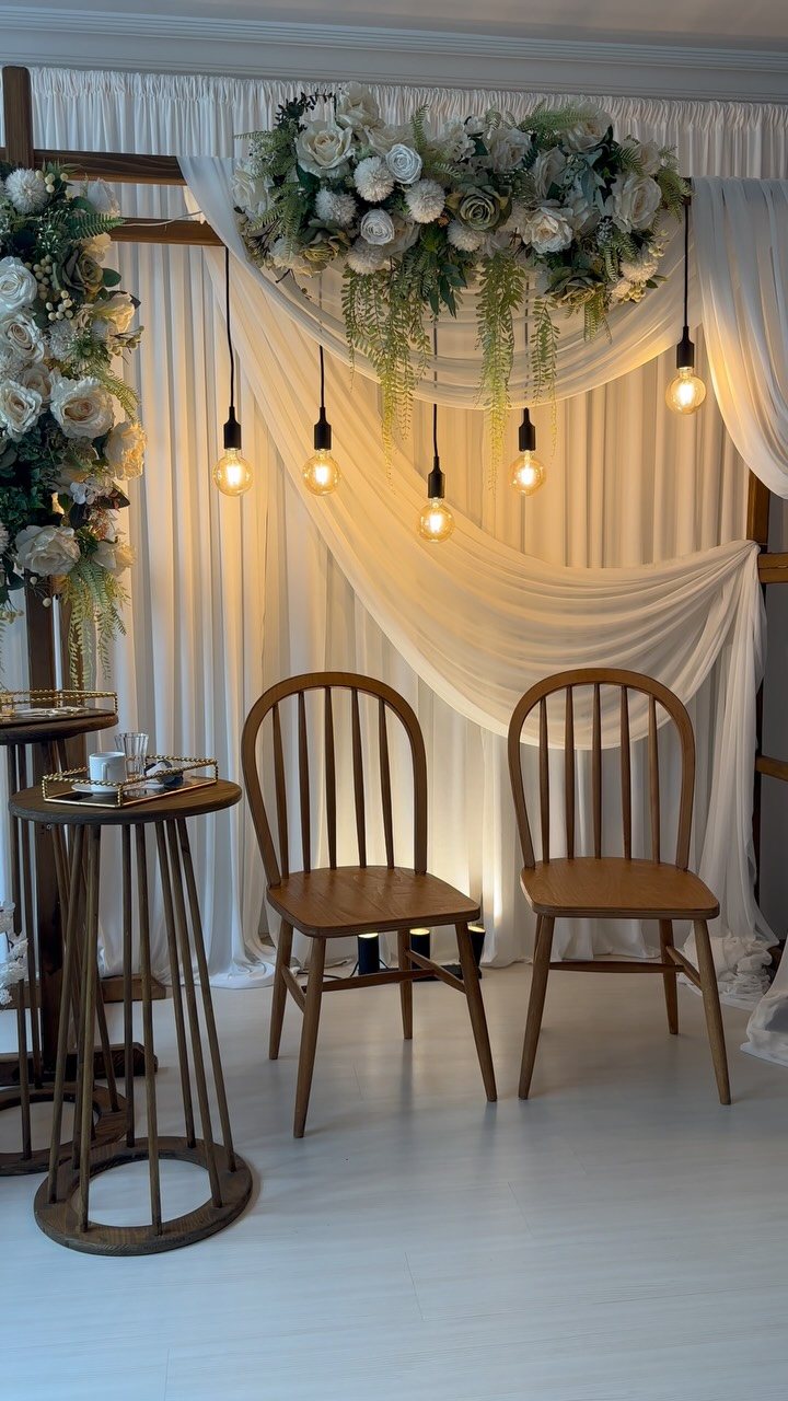 White draped fabric backdrop with hanging light bulbs and a large floral arrangement above wooden chairs.