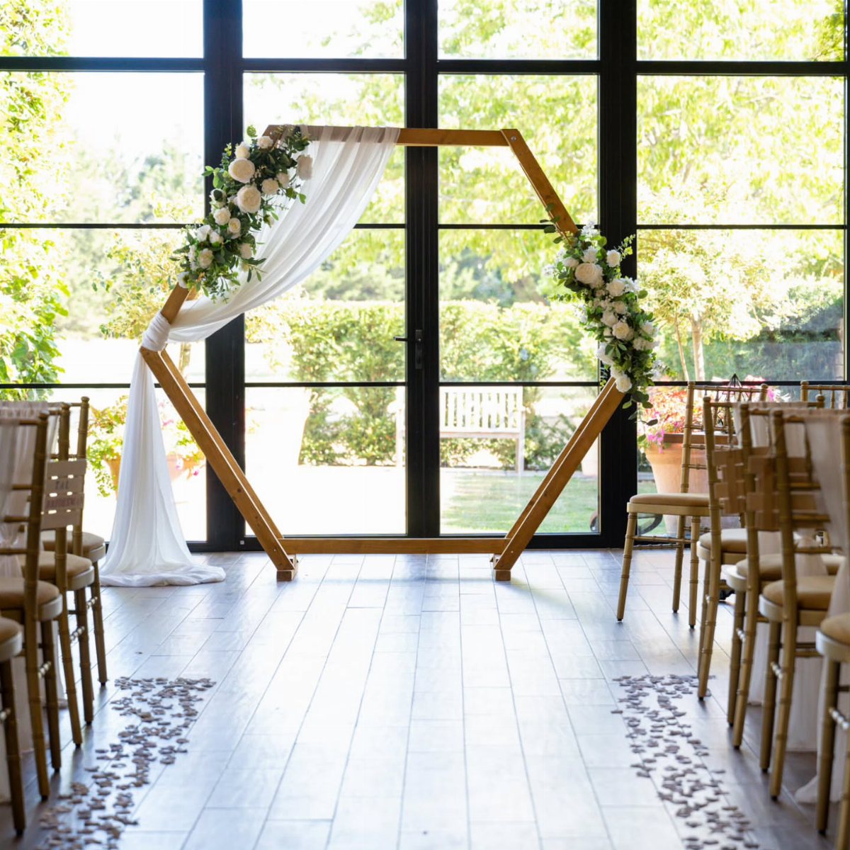 Wooden hexagonal wedding arch with white floral arrangements and draped fabric in a bright indoor setting.
