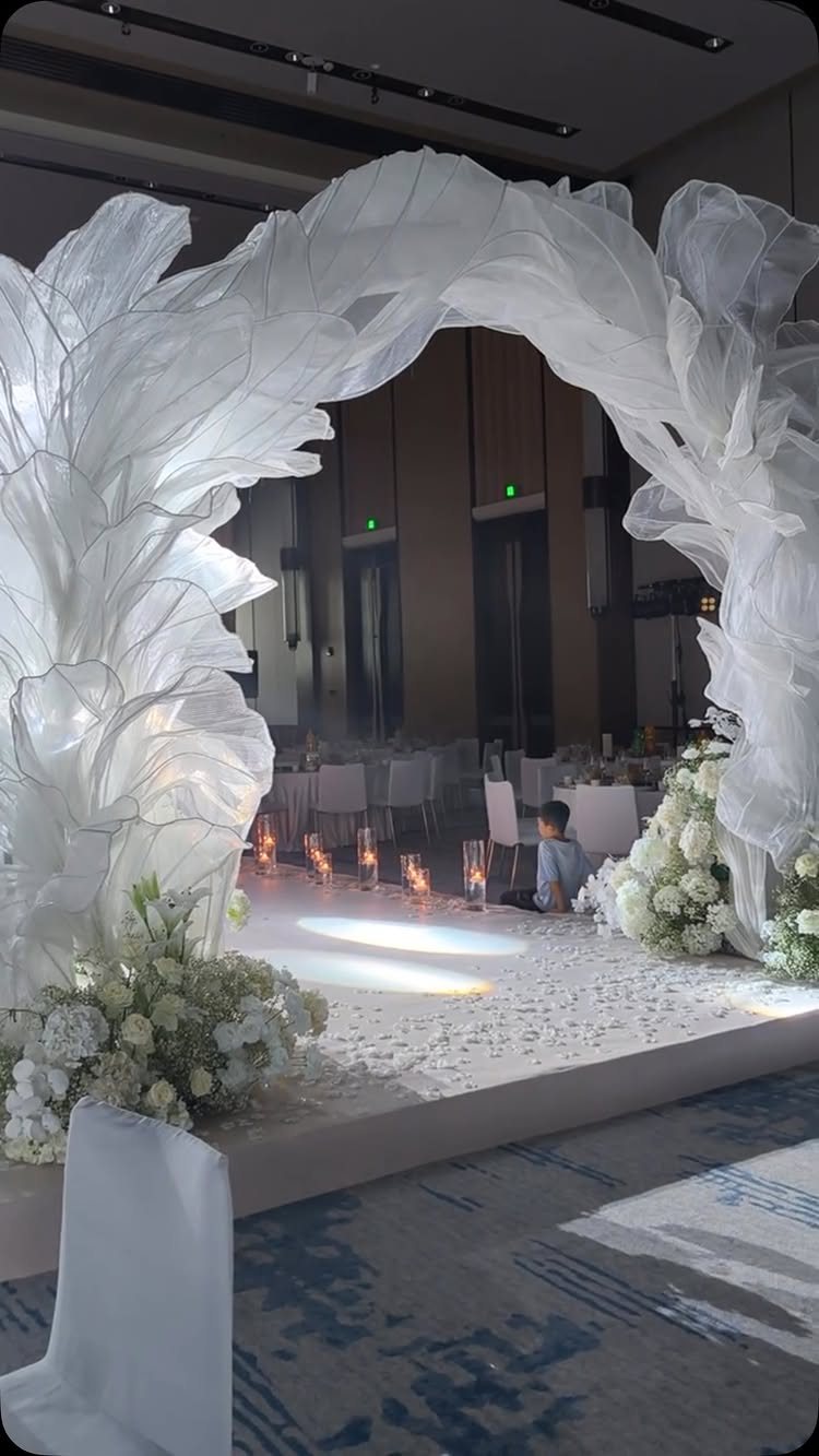 Large white sculptural fabric wedding arch with floral arrangements in a dimly lit indoor ballroom.