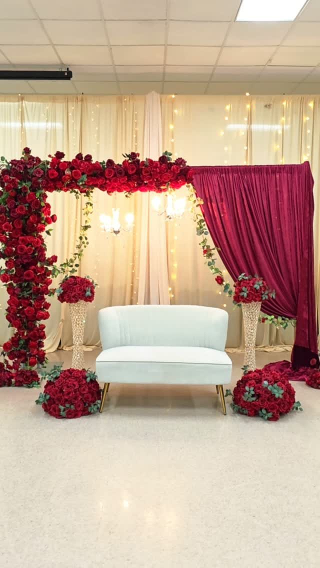Red rose arch with burgundy velvet drapery and a white loveseat against twinkling lights.