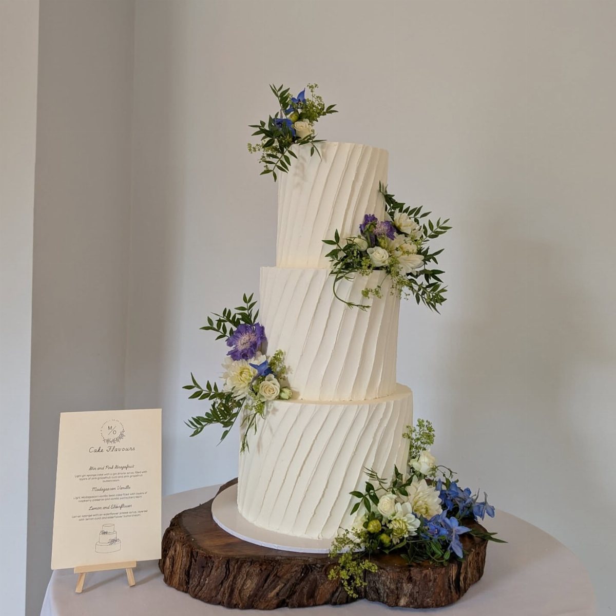 Three-tier white wedding cake with textured buttercream icing and blue and white floral arrangements.